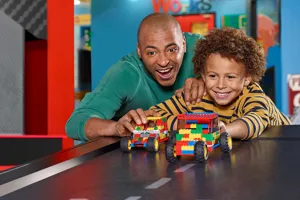 An excited adult and child racing colorful LEGO cars at the LEGOLAND Discovery Center's creative workshop.