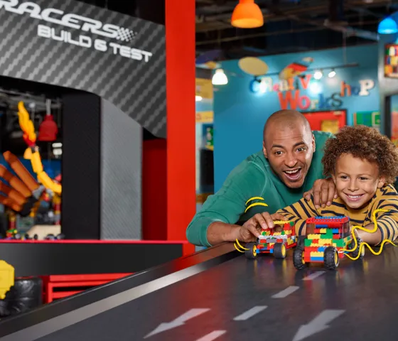A dad and his son are building a LEGO race car together at LEGOLAND Discovery Center Bay Area. The dad is helping his son assemble the car using colorful LEGO bricks, while both focus on the creative process. The scene is set within a bright, interactive area of the center, with other LEGO structures and activities visible in the background. The father and son share a moment of excitement and teamwork as they work on their race car creation.